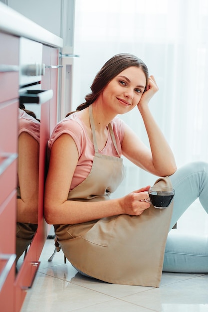 Jolie femme au foyer portant un tablier dans la cuisine reposant sur le sol avec une tasse de café