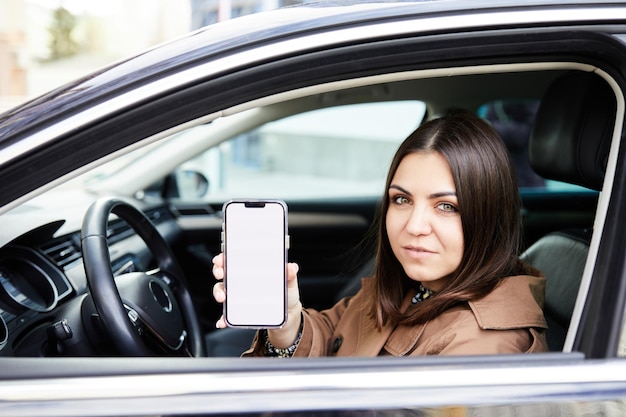Jolie femme au volant d'une voiture, elle montre un écran de téléphone vide et sourit