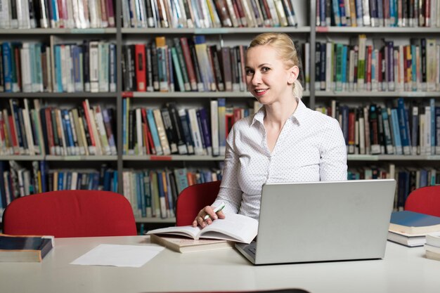 Jolie femme aux cheveux blonds assis à un bureau dans la bibliothèque ordinateur portable et organiseur sur la table en regardant l'écran un concept d'étude de livres flous à l'arrière