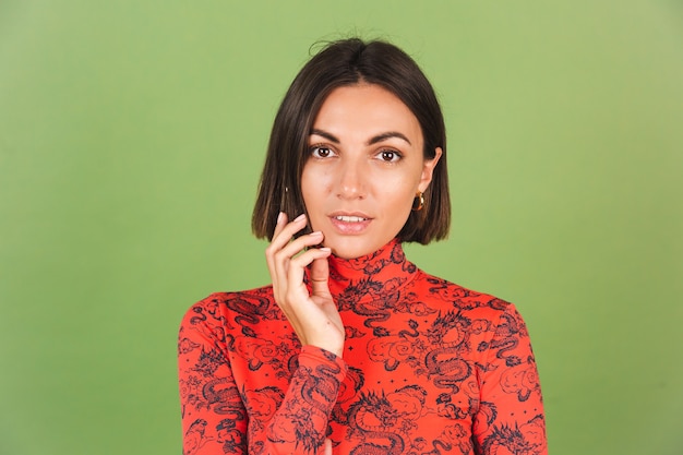 Jolie femme aux cheveux courts avec des boucles d'oreilles dorées légères, un chemisier imprimé de dragon de Chine rouge sur des émotions positives vertes, un sourire confiant