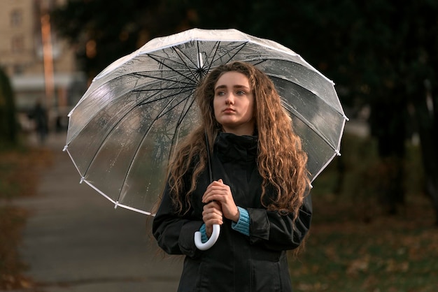 Une jolie femme aux cheveux longs se promène dans le parc d'automne par temps de pluie sous un parapluie transparent