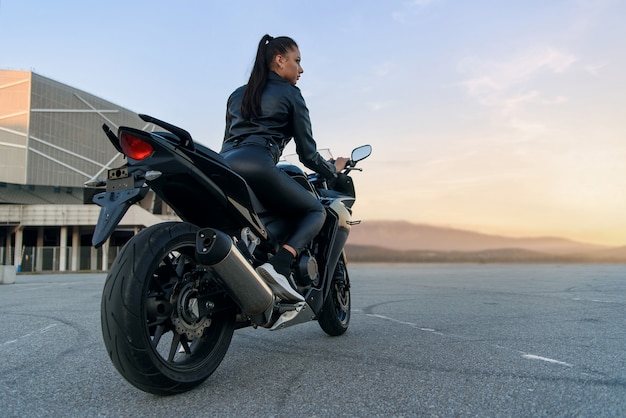Jolie femme aux cheveux longs en veste et pantalon en cuir noir sur un parking extérieur avec une moto de sport élégante au coucher du soleil.