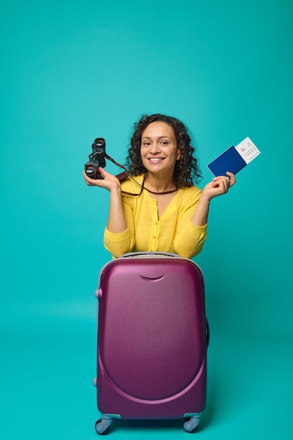 Jolie femme aux cheveux noirs bouclés heureux sourit à pleines dents en regardant la caméra, tenant des jumelles et un passeport avec carte d'embarquement et billet, posant avec une valise sur fond bleu. Copier l'espace publicitaire