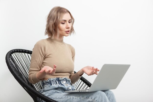 Jolie femme blonde a un appel vidéo ou une conférence sur un ordinateur portable tout en étant assis sur une chaise sur fond de mur blanc à la maison Concept de travail à distance indépendant