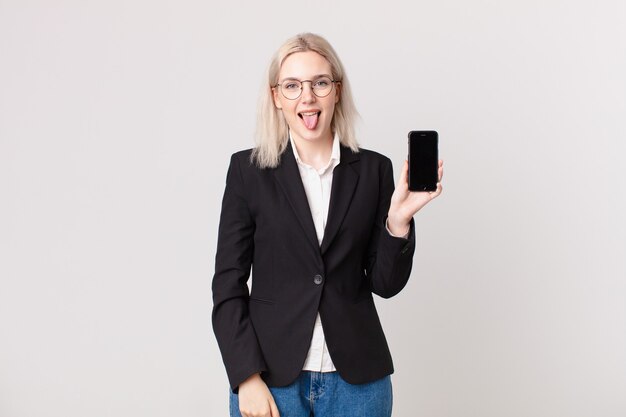 Jolie femme blonde avec une attitude joyeuse et rebelle, plaisantant et tirant la langue et tenant un téléphone portable