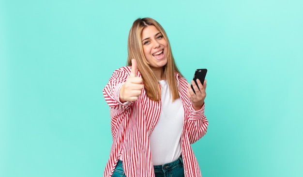 Jolie femme blonde bien roulée se sentant fière, souriante positivement avec les pouces vers le haut et tenant un téléphone intelligent