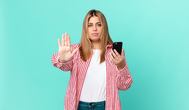 Jolie femme blonde bien roulée à la sérieuse montrant la paume ouverte faisant un geste d'arrêt et tenant un téléphone intelligent