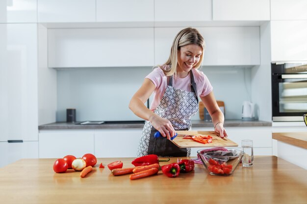 Jolie femme blonde caucasienne souriante digne en tablier debout dans la cuisine et mettre des légumes hachés dans un bol.