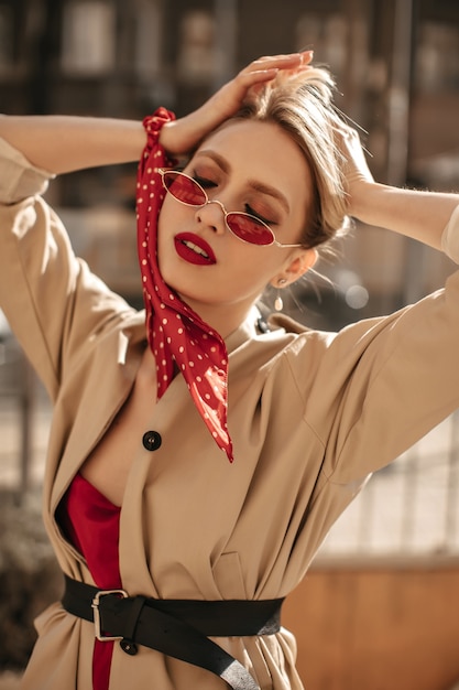 Jolie femme blonde charmante aux lèvres rouges pose à l'extérieur. Une femme séduisante dans des lunettes de soleil colorées et un trench-coat beige touche les cheveux