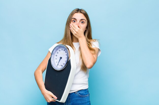 Jolie femme blonde couvrant la bouche avec les mains avec une expression choquée et surprise, gardant un secret ou disant oups