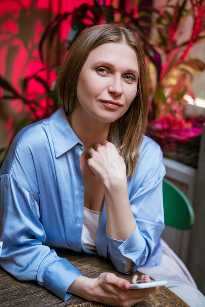 Une jolie femme blonde dans une chemise bleue à une table dans un café avec un téléphone à la main se penche sur la caméra