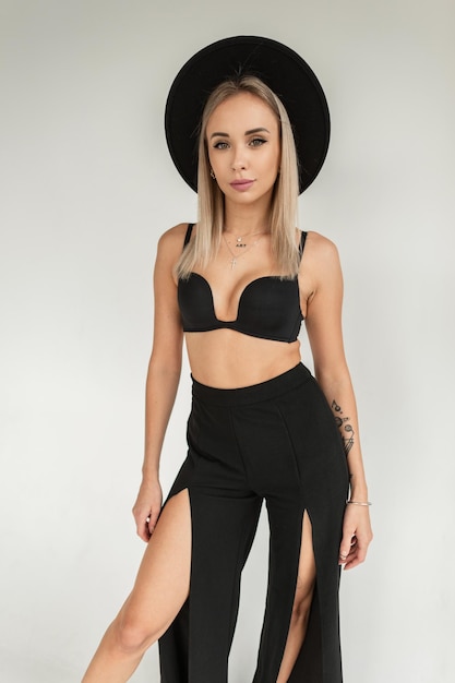 Jolie femme blonde élégante et chaude avec un chapeau noir vintage en soutien-gorge de mode et un pantalon sexy près d'un mur blanc à l'intérieur