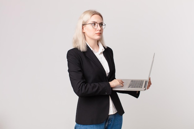 Jolie femme blonde haussant les épaules, se sentant confuse et incertaine et tenant un ordinateur portable