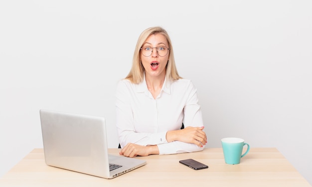 Jolie femme blonde jeune femme blonde ayant l'air très choquée ou surprise et travaillant avec un ordinateur portable