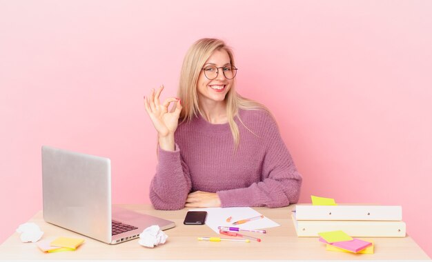 Jolie femme blonde jeune femme blonde se sentant heureuse, montrant son approbation avec un geste correct et travaillant avec un ordinateur portable