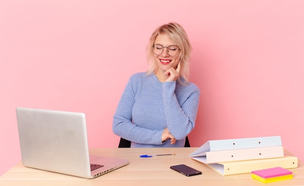 Jolie femme blonde jeune jolie femme souriante avec une expression heureuse et confiante avec la main sur le menton. concept de bureau d'espace de travail