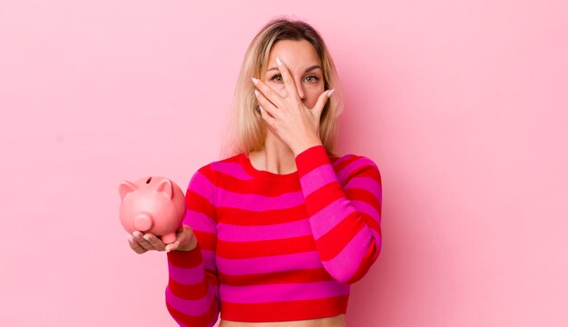 Jolie femme blonde à la peur choquée ou terrifiée couvrant le visage avec le concept de tirelire à la main