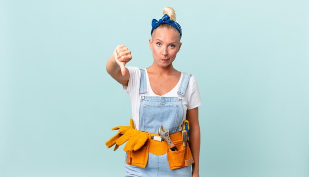 Jolie femme blonde se sentant croisée, montrant les pouces vers le bas et réparant le concept de maison