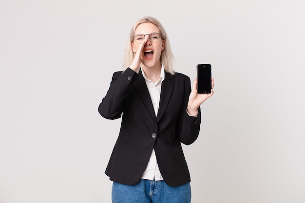 Jolie femme blonde se sentant heureuse, donnant un grand cri avec les mains à côté de la bouche et tenant un téléphone portable