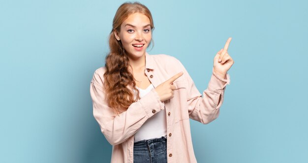 Jolie femme blonde se sentant joyeuse et surprise, souriant avec une expression choquée et pointant vers le côté