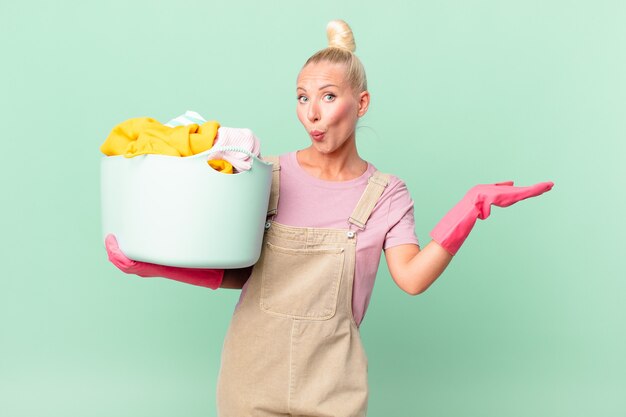 Jolie femme blonde semblant surprise et choquée, avec la mâchoire tombée tenant un objet concept de lavage de vêtements