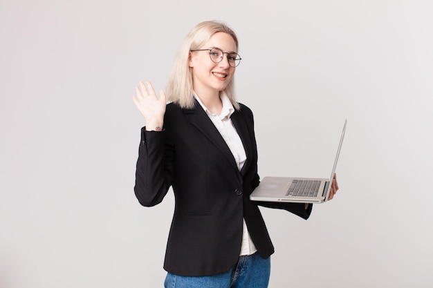 Jolie femme blonde souriant joyeusement, agitant la main, vous accueillant et vous saluant et tenant un ordinateur portable