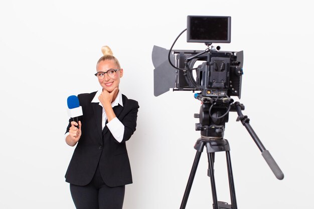 Jolie femme blonde souriante avec une expression heureuse et confiante avec la main sur le menton et tenant un micro. concept de présentateur