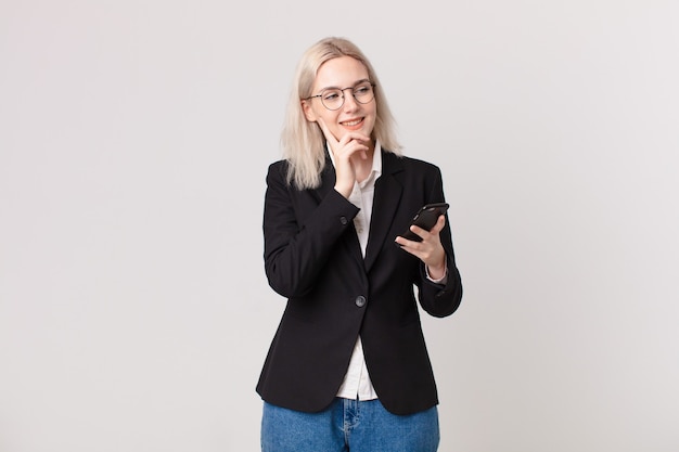 Jolie femme blonde souriante avec une expression heureuse et confiante avec la main sur le menton et tenant un téléphone portable