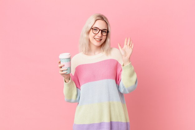 Jolie femme blonde souriante joyeusement, agitant la main, vous accueillant et vous saluant. concept de café