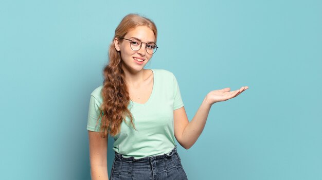 Jolie femme blonde souriante joyeusement, se sentant heureuse et montrant un concept dans l'espace de copie avec la paume de la main