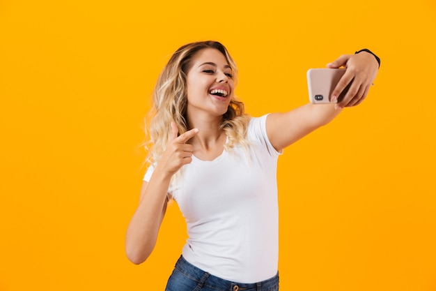 Jolie femme blonde souriante et prenant selfie photo sur téléphone mobile, isolé sur mur jaune