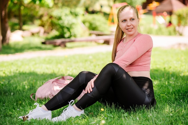 Jolie femme blonde en vêtements de sport et sac de sport assis et se reposant sur la pelouse dans le parc