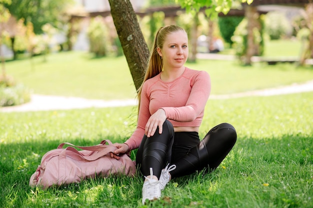 Jolie femme blonde en vêtements de sport et sac de sport assis et se reposant sur la pelouse dans le parc