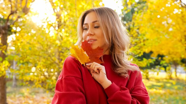 Jolie femme blonde vêtue d'une robe chaude rouge avec des feuilles d'érable jaunes posant pour la caméra dans un parc d'automne ensoleillé en gros plan