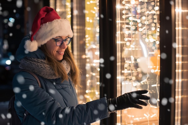 Jolie femme en bonnet de Noel regardant et rêvant dans la vitrine illuminée