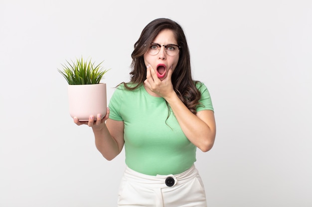 Jolie femme avec la bouche et les yeux grands ouverts et la main sur le menton et tenant une plante décorative