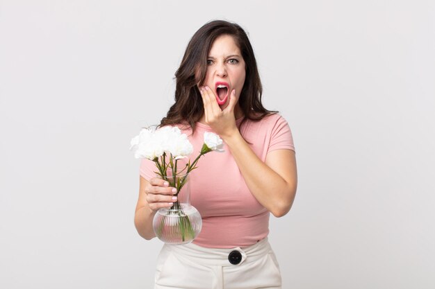 Jolie femme avec la bouche et les yeux grands ouverts et la main sur le menton et tenant un pot de fleurs décoratif