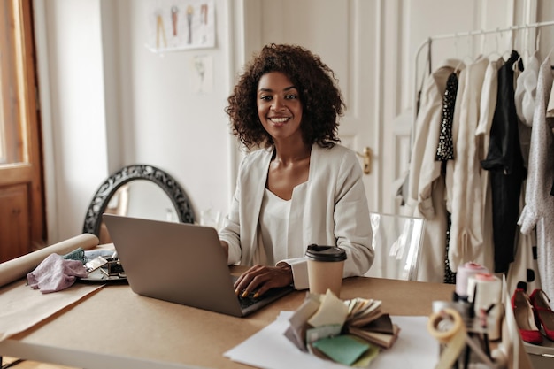 Jolie femme brune bouclée à la peau foncée en veste beige sourit largement assis près du bureau et travaille dans un ordinateur portable dans un bureau de designer confortable