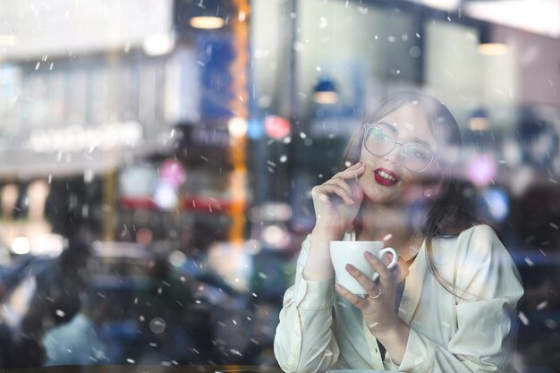 Jolie femme brune avec du rouge à lèvres rouge garde au chaud avec une tasse de café chaud dans un restaurant. Photo à travers la fenêtre par temps de neige. Espace pour le texte