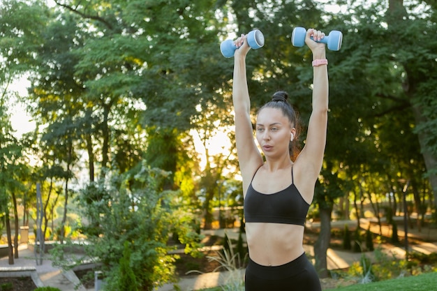 Jolie femme brune en forme de caucasienne entraînant les muscles des épaules avec des haltères dans ses mains dans le parc au lever du soleil