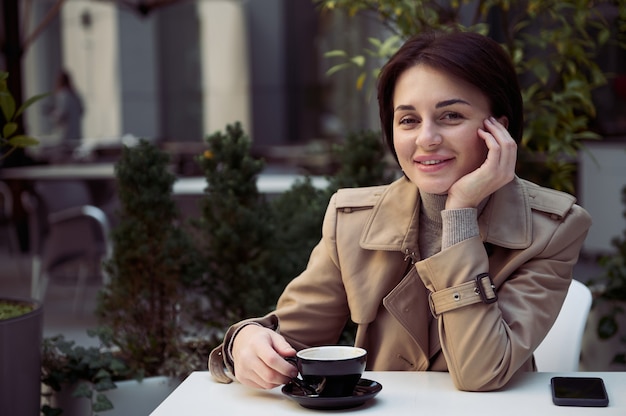 Jolie femme brune heureuse dans un trench-coat beige en cuir écologique élégant assis à une table dans une cafétéria en plein air et regardant la caméra en profitant de sa pause-café