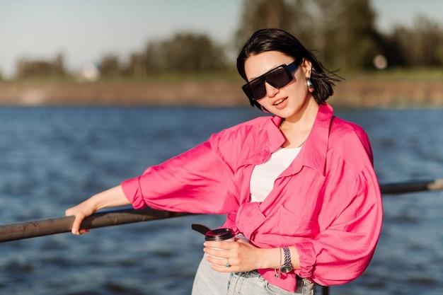 Jolie femme brune à lunettes de soleil avec une tasse de café sur la jetée au bord du lac