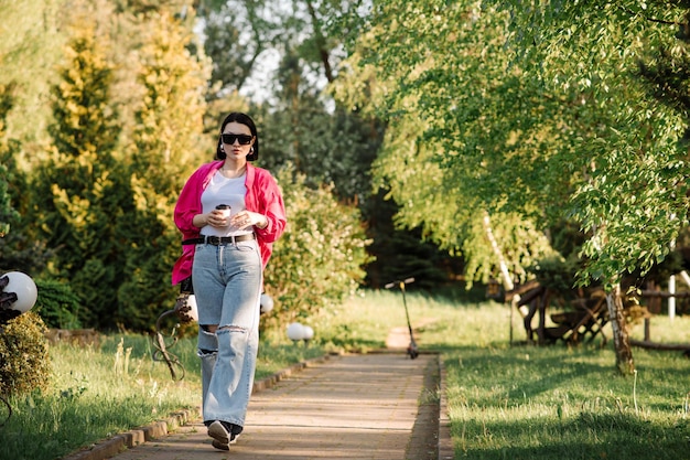 Jolie femme brune à lunettes de soleil avec une tasse de café marchant dans le parc