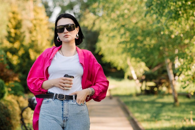 Jolie femme brune à lunettes de soleil avec une tasse de café marchant dans le parc