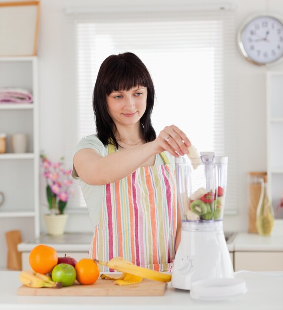 Jolie femme brune mettant des légumes dans un mélangeur en position debout