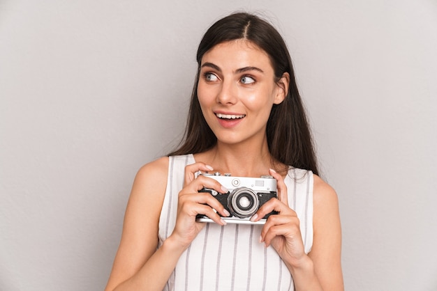 Jolie femme brune portant une robe souriante et tenant une photocaméra rétro isolée sur un mur gris