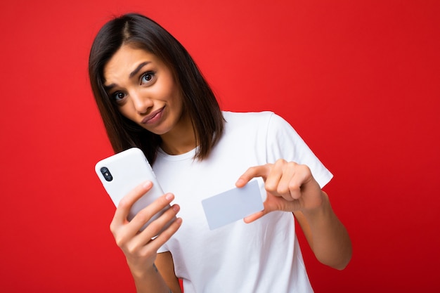 Jolie femme brune portant un t-shirt blanc élégant de tous les jours isolé sur un mur de fond rouge tenant et utilisant un téléphone et une carte de crédit effectuant le paiement en regardant la caméra et en ayant des questions