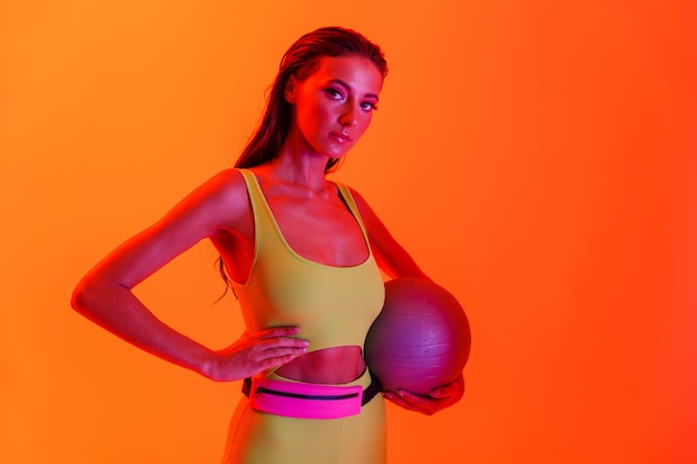 Jolie femme brune portant des vêtements de sport ajustés tenant un ballon de gymnastique tout en faisant de l'exercice isolé sur un mur de néon orange