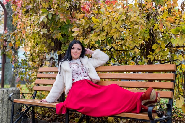 Jolie femme brune en robe rouge assis sur un banc en automne parc