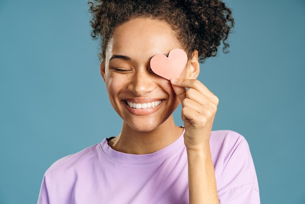 Jolie femme brune souriante tenant un coeur rose près de son œil et souriant tout en soutenant une autre femme. Studio intérieur tourné isolé sur fond rose